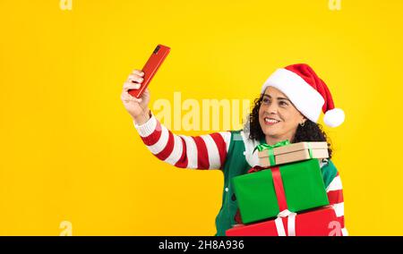 Portrait d'une femme latine adulte d'âge moyen tenant un téléphone mobile prenant un selfie photo dans un concept de noël sur fond jaune Banque D'Images