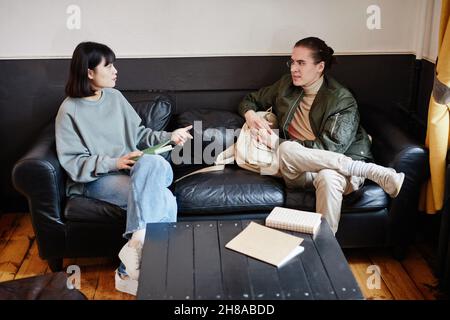 Deux amis de l'école assis sur un canapé et discutant de leurs devoirs dans l'équipe dans la salle Banque D'Images
