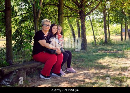 Vue sur toute la longueur d'une famille heureuse assise sur un banc en bois dans un parc sur un fond d'arbres verts Banque D'Images