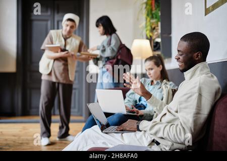 Des étudiants africains heureux agitant et souriant à la caméra tout en étant assis sur le canapé et en parlant en ligne sur ordinateur portable avec d'autres étudiants dans le bakground Banque D'Images