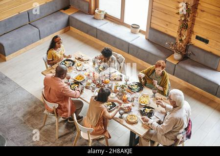 Famille interraciale de six personnes qui mangent de la nourriture de fête maison tout en étant assis près d'une table dans la salle à manger avec des murs en bois et de grandes fenêtres Banque D'Images