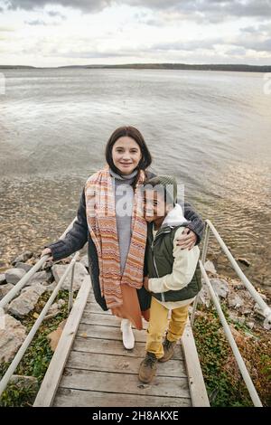 Bonne jeune femme caucasienne et son mignon fils interracial en tenue décontractée chaude debout contre le lac le week-end d'automne Banque D'Images