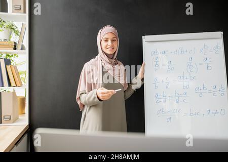 Joyeux jeune professeur de hijab debout par tableau blanc avec formule algébrique et équations tout en les expliquant devant l'ordinateur Banque D'Images