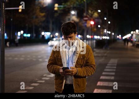 Messages masculins sur les médias sociaux par téléphone mobile en traversant la route en ville le soir Banque D'Images