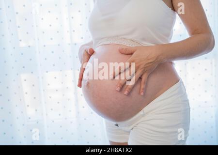 Crop anonyme femme enceinte frottant ventre près de la fenêtre Banque D'Images