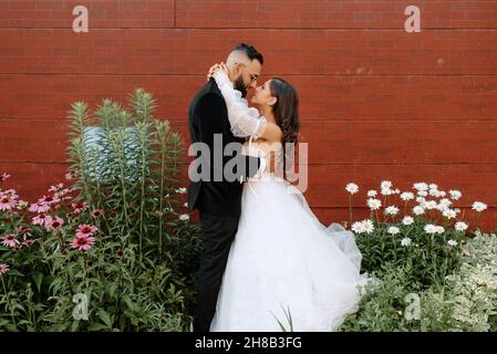 Mariée et marié embrassant contre le mur de brique et les fleurs Banque D'Images