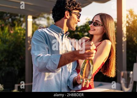 Un couple aimant ouvre une bouteille de champagne à côté des amis assis à la table pour célébrer la fête d'engagement Banque D'Images