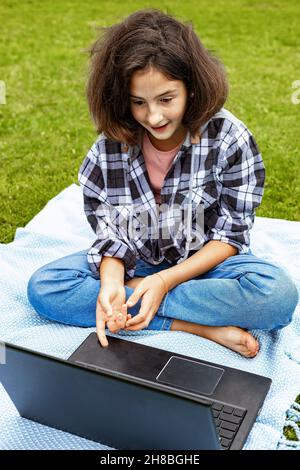 Une jeune fille adorable assise sur l'herbe et travaillant sur un ordinateur portable. Une écolière regarde une classe Internet et étudie à distance. La fille utilise le sans fil Banque D'Images