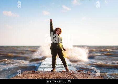 Une femme heureuse en surpoids a joyeusement jeté sa main, debout sur la jetée au bord de la mer.Ciel et océan en arrière-plan.Le concept de psycholo positif Banque D'Images