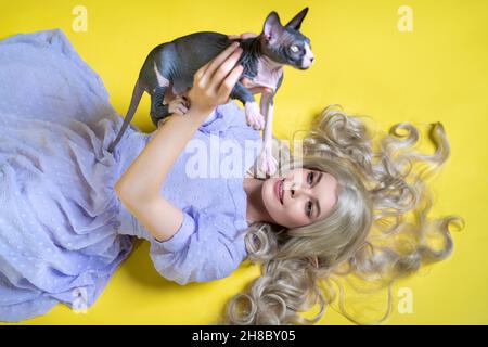 Cosplayer elf jeune femme en robe bleue couché sur fond jaune, tient Sphinx chat au-dessus de sa tête, sourit et regarde Banque D'Images