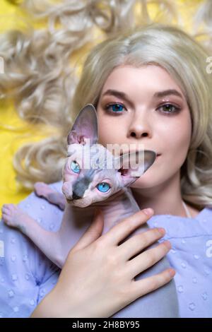 Portrait de jeune femme blonde cosplay elf en robe bleue avec les yeux de différentes couleurs couchés sur fond jaune, tenant sur sa poitrine Sphinx chat Banque D'Images