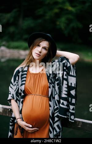 La femme enceinte dans un chapeau s'appuie sur la rampe d'un quai sur le lac.Portrait Banque D'Images