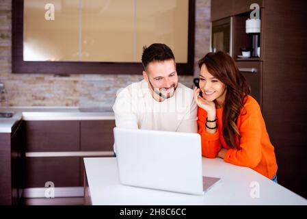 Sourire heureux couple debout dans la cuisine à la maison et à l'aide d'un ordinateur portable. Banque D'Images