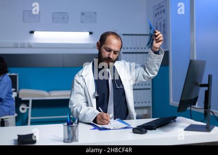 Spécialiste en imagerie radiographique pour examen tout en prenant des notes sur le papier de contrôle.Un médecin spécialisé rédigeant le diagnostic et les résultats après une analyse radiographique pour un traitement médical de nuit. Banque D'Images