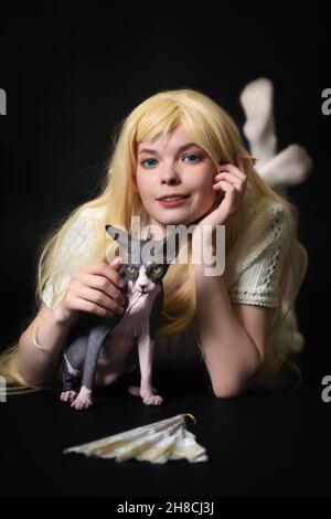 Jeune femme cosplayer elf en robe tricotée blanche, avec de longs cheveux blonds couchés sur le ventre sur fond noir, souriant et regardant l'appareil photo Banque D'Images