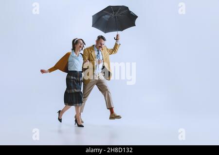 Jeune famille heureux couple, homme et femme dans des vêtements de style rétro, mode des années 70, années 60 marche avec parapluie isolé sur fond blanc studio Banque D'Images