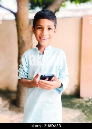 Portrait d'un garçon sud-asiatique debout, souriant et tenant un téléphone portable dans ses mains à l'extérieur Banque D'Images