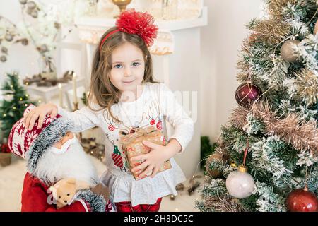 Adorable fille caucasienne portant une tenue festive et tenant un cadeau entouré par le décor des vacances d'hiver avec une grande figurine de Santa et des jouets Banque D'Images