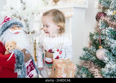 Adorable petite fille blonde portant un pyjama festif tout en souriant et jouant avec la multitude de jouets entourant l'arbre de Noël décoré Banque D'Images