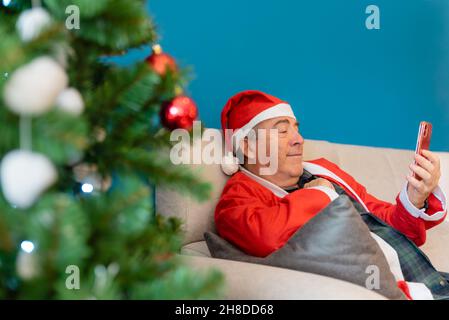 Grand-père habillé comme le Père Noël regarde le téléphone mobile tout en étant assis sur le canapé avec un fond bleu Banque D'Images