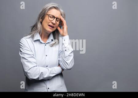Femme d'affaires séniée, sénior et sénior, à cheveux gris, debout, isolée sur le front, avec les yeux fermés, femme aînée chef de la direction, se sent mal de tête, souffre de migraine, de l'épuisement professionnel Banque D'Images