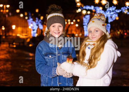 Drôle couple d'enfants, fille et garçon, tenir la lanterne dans leurs mains contre le fond des lumières de noël dehors, dans les vêtements chauds d'hiver et les chapeaux tricotés.Concept de Noël et d'enfance, extérieur Banque D'Images