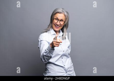 Hé vous.Souriante positive heureuse femme d'affaires à cheveux gris regarde la caméra et pointe l'index vers vous, femme mûre en tenue habillée isolée sur gris, vous choisit Banque D'Images