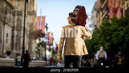Latino homme marchant dans une rue animée avec une guitare sur son épaule de derrière Banque D'Images