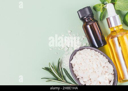 Set d'aromathérapie avec des bouteilles d'huile essentielle naturelle à côté d'un bol de sel de bain de mer et des branches d'herbes fraîches romarin et menthe sur fond vert Banque D'Images