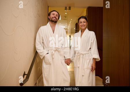 Beau couple affectueux, portant des peignoirs blancs, profitant de leur lune de miel ensemble dans un luxueux spa, en tenant les mains pendant qu'ils descendent le Banque D'Images