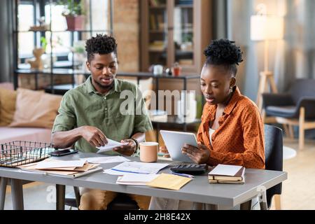 Jeune couple afro-américain concentré assis à table avec des factures et analysant les dépenses mensuelles à la maison Banque D'Images