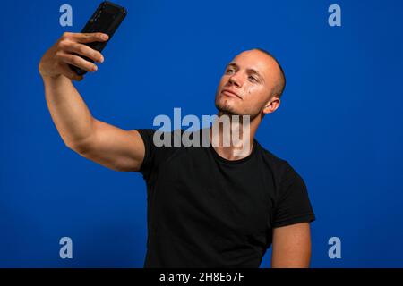 Beau jeune homme caucasien avec des cheveux blonds et des yeux bleus prenant un selfie isolé sur fond bleu studio Banque D'Images