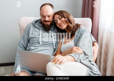 Jeune beau couple marié sont assis sur un fauteuil à l'intérieur de la maison à l'aide d'un ordinateur portable.Bonne femme souriante et homme faire des achats en ligne via Internet sur Banque D'Images