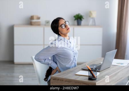 Fatiguée bouleversée jeune femme d'affaires indienne souffrant de douleurs dorsales, spasme musculaire au travail avec ordinateur portable Banque D'Images