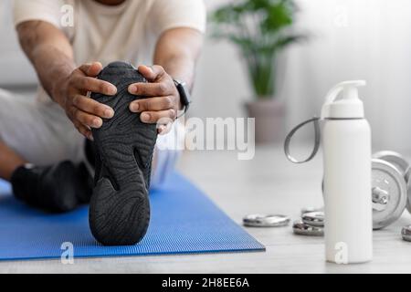 Jeune afro-américain court faisant un étirement de jambe sur le tapis sur le sol avec des haltères et une bouteille d'eau Banque D'Images