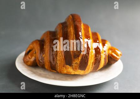 Un croissant français fraîchement préparé avec garniture au chocolat se trouve sur une soucoupe blanche sur une table grise.Concept de boulangerie traditionnelle. Banque D'Images