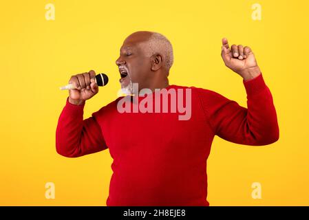 Cool senior Black man chantant la chanson, en utilisant le microphone, en exécutant le karaoké sur fond orange studio Banque D'Images