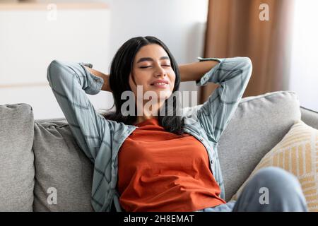 Jeune femme arabe détendue, penchée sur un canapé, se reposant avec les mains derrière la tête sur un canapé confortable, fermant les yeux Banque D'Images