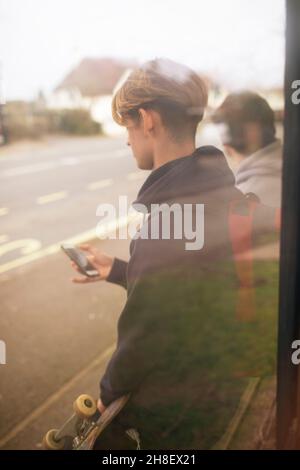 Adolescent avec smartphone et skateboard à l'arrêt de bus Banque D'Images