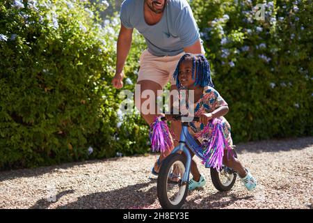 Père aidant la petite fille à faire du vélo dans une allée ensoleillée Banque D'Images