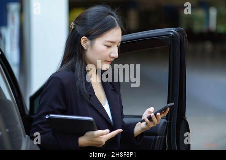 Femme d'affaires asiatique occupée à sortir de son taxi tout en utilisant un message texte pour smartphone à son assistant. Banque D'Images