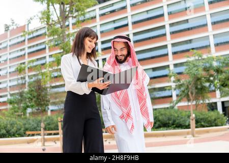 D'en dessous de la femme de direction dans des vêtements élégants montrant le contrat dans un dossier à un client arabe gai mâle à keffiyeh lors d'une réunion d'affaires dans la rue de la ville moderne Banque D'Images