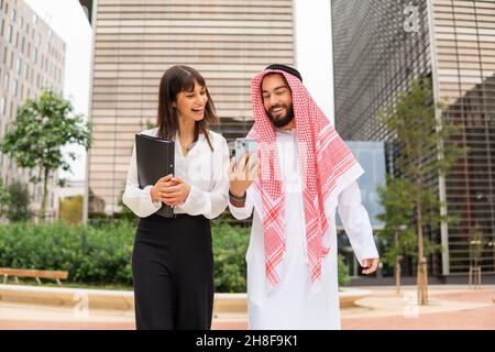 Un homme arabe heureux, vêché de vêtements traditionnels, montrant un téléphone portable à une femme gestionnaire optimiste tout en marchant dans la rue contre des gratte-ciel lors d'une réunion d'affaires dans le centre-ville Banque D'Images