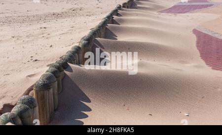 Le sable qui se déplace sur la plage souffle sur des rôles en bois peu élevés sur le chemin de promenade dans des monticules de pieux de sable de grain pendant la saison des vents forts. Banque D'Images