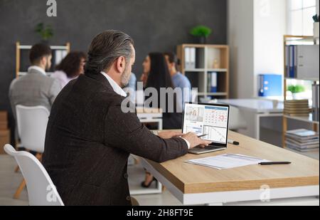 Homme d'affaires analyse son travail en regardant le programme avec l'interface affichée sur l'écran d'ordinateur portable. Banque D'Images