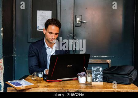 Berlin, Allemagne. Jeune adulte et professionnel gay travaillant avec son ordinateur portable et son smartphone à l'intérieur d'un café pour aider ses clients avec leur marketing en ligne et seo. Banque D'Images