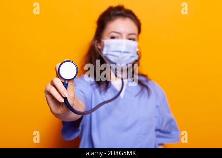 Gros plan de l'infirmière avec masque facial montrant le stéthoscope sur la caméra en studio.Assistant médical avec protection du coronavirus ayant uniforme et équipement de santé utilisé pour le contrôle. Banque D'Images