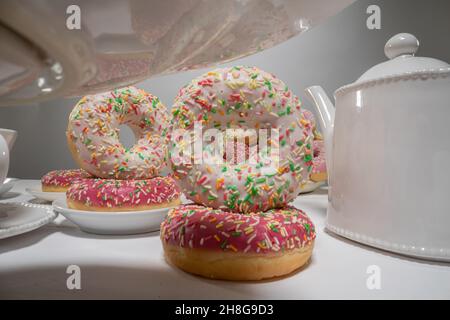 Beignets multicolores avec arrosage et glaçage.Table pleine de beignets, tasses, café et verseuse.Effet de parallaxe. Banque D'Images