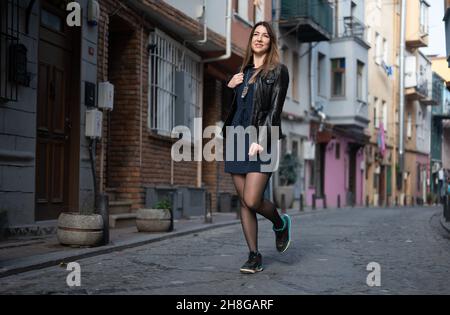 Grande fille attrayante marche le long d'une rue étroite de la ville. La jeune femme porte une veste en cuir et une robe courte. Regardez dans la distance. Pleine dbo Banque D'Images