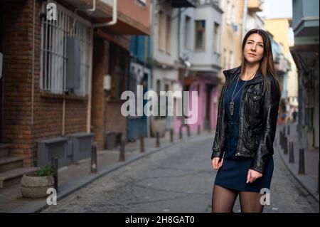 Grande femme attrayante marche le long de la rue étroite de la ville. La jeune femme porte une veste en cuir et une robe courte. Regardez la distance Banque D'Images